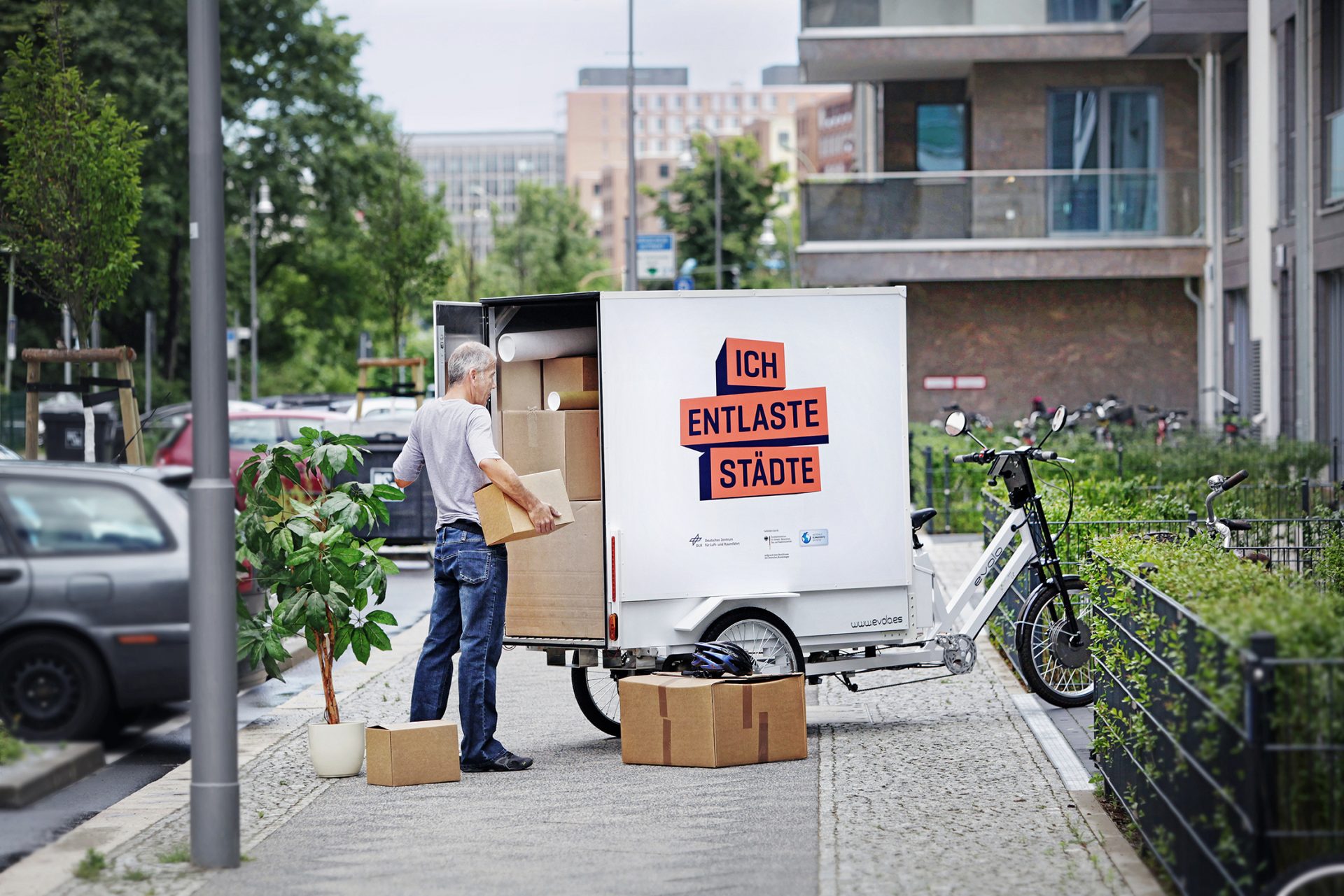 Sur le trottoir, un homme charge des cartons de déménagement sur un grand vélo-cargo. Sur la roue, il y a 