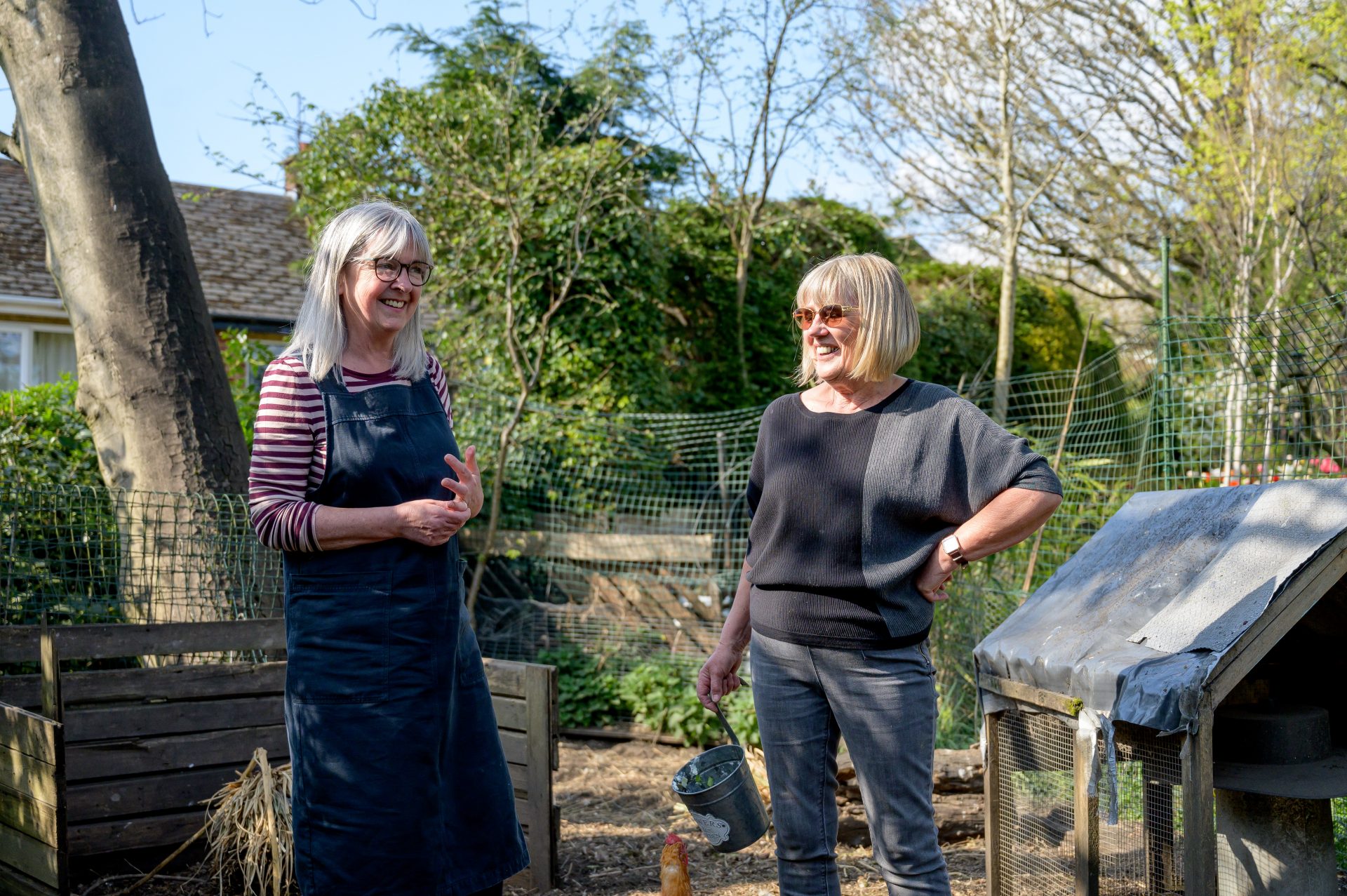 Deux femmes âgées se tiennent dans un jardin au soleil et discutent en riant.